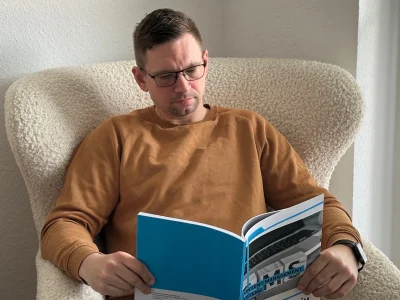 Ein Mann mit Brille sitzt in einem gemütlichen, hellen Polstersessel und liest aufmerksam in einem blauen Studienheft der IU mit dem Titel „Content Management Systeme"