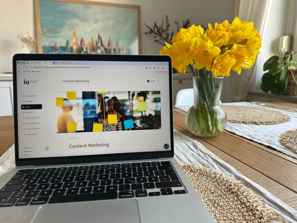 Ein geöffneter Laptop auf einem Holztisch zeigt das Online-Lernportal der IU mit dem Kurs „Content-Marketing“. Daneben steht eine Glasvase mit leuchtend gelben Narzissen.