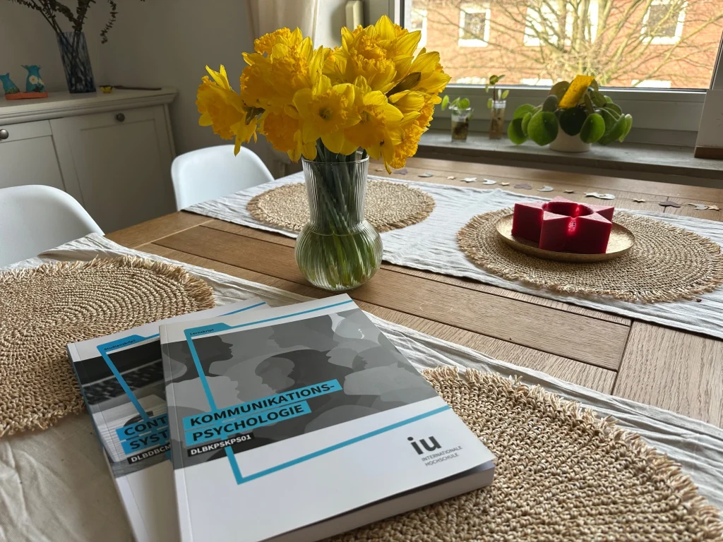 Zwei Studienhefte der IU Internationalen Hochschule mit den Titeln „Kommunikationspsychologie“ und „Content Management Systeme“ liegen auf einem gedeckten Tisch neben gelben Frühlingsblumen.