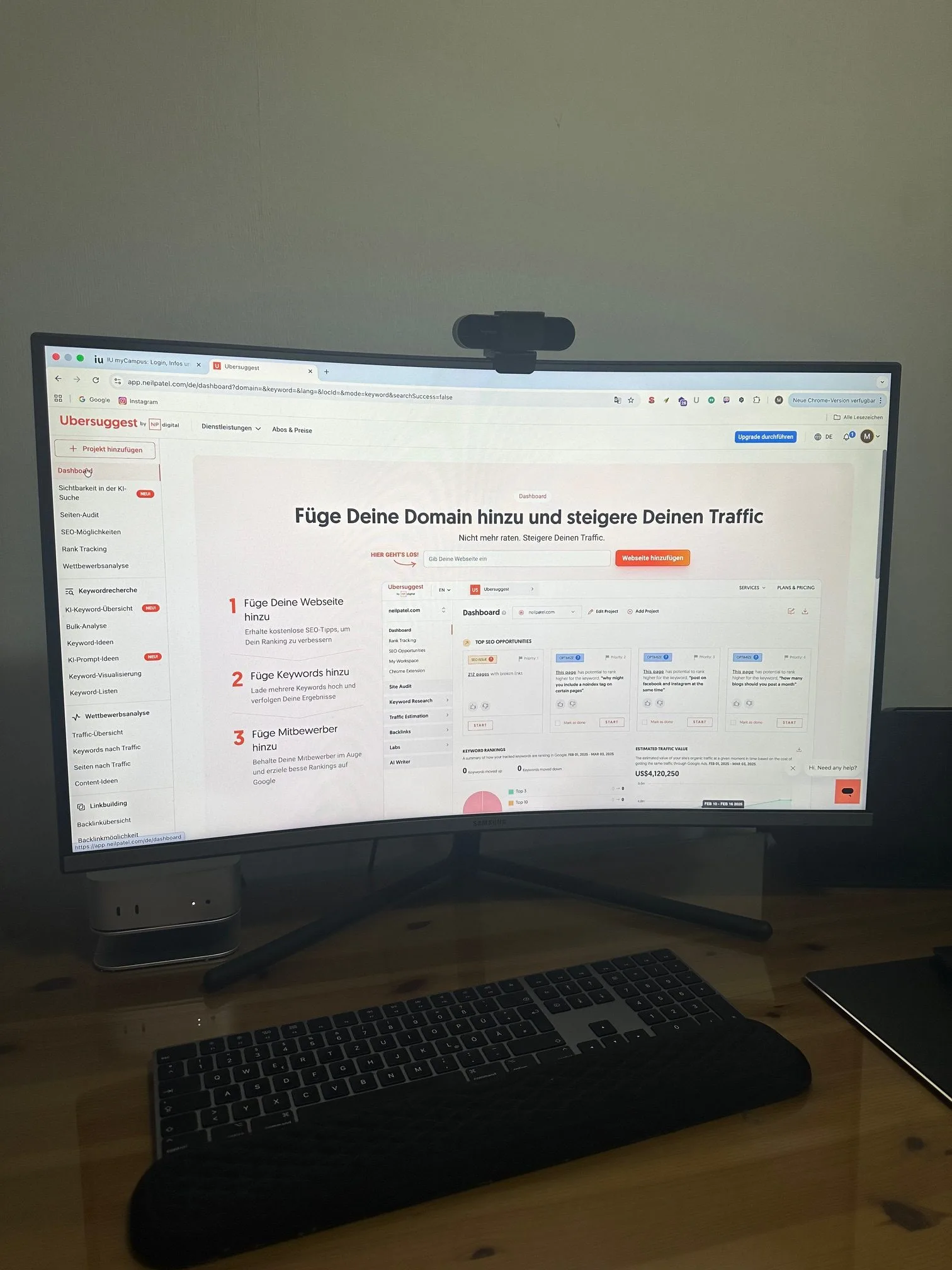 Ein moderner Home-Office-Arbeitsplatz mit einem Curved-Monitor von Samsung, auf dem das SEO-Tool „Ubersuggest“ von Neil Patel geöffnet ist. Die Webseite zeigt das Dashboard mit der Aufforderung „Füge Deine Domain hinzu und steigere Deinen Traffic“. Vor dem Monitor liegen eine flache, dunkle Tastatur und ein Trackpad auf einem hellen Holzschreibtisch.