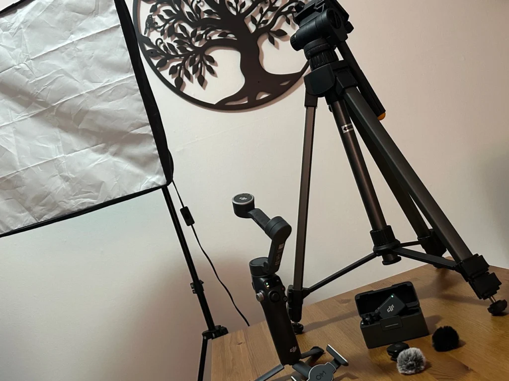 Das Bild zeigt eine günstige Social-Media-Technikausstattung. Ein Gimbel, Mikrofone, ein altes Licht und ein altes Stativ auf einem Holztisch liegen.